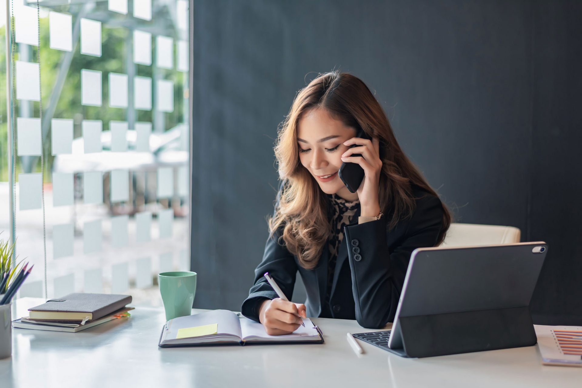 woman taking notes on pad while talking on cellphone in office environment